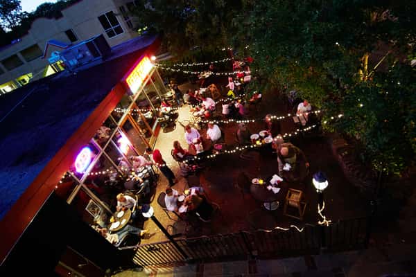 Bustling outdoor restaurant patio at night with diners enjoying meals under string lights and a glowing neon sign.