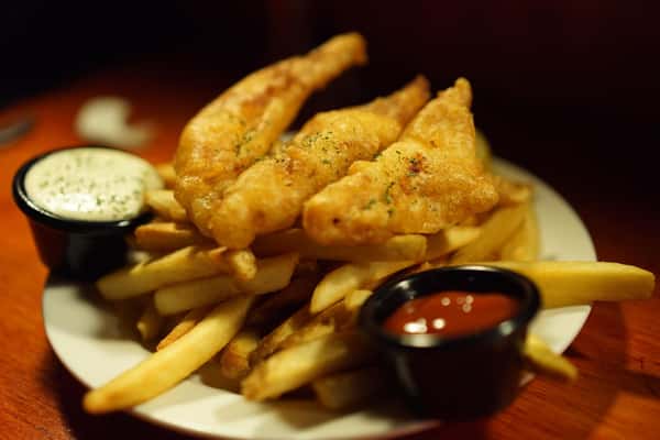 Golden beer-battered fish fillets served over crispy French fries with tartar sauce and ketchup—classic fish and chips plated in a casual restaurant setting.
