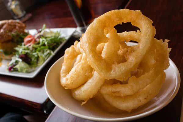Crispy beer-battered onion rings stacked in a bowl, served as a classic pub appetizer with a blurred burger and salad in the background.