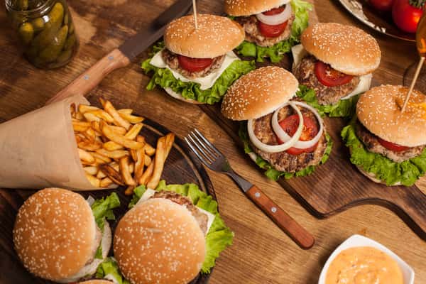 A wooden table topped with cheeseburgers, fresh lettuce, tomato, onions, a cone of fries, pickles, and a bowl of dipping sauce.