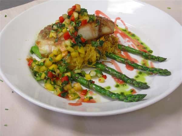 Seared fish fillet served over a bed of spaghetti squash and grilled asparagus, topped with a vibrant mango salsa and drizzled sauce.