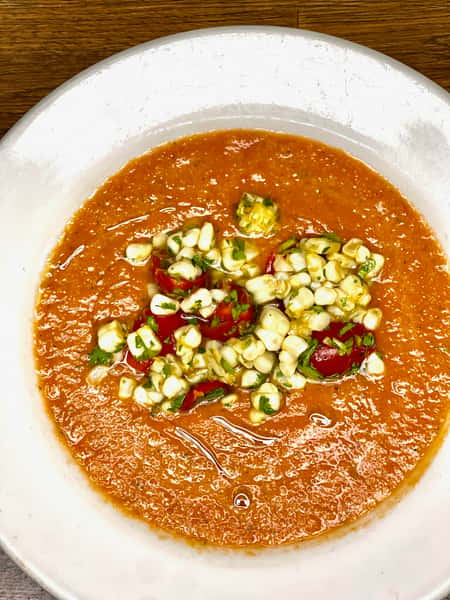 Chilled tomato gazpacho topped with fresh corn, cherry tomatoes, and herbs served in a white bowl.