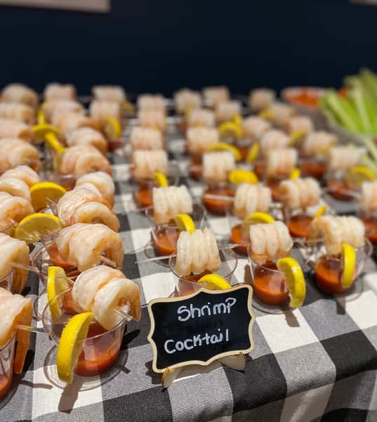 Individual shrimp cocktail cups with lemon wedges and cocktail sauce arranged on a checkered tablecloth, perfect for upscale catering or seafood-themed parties.