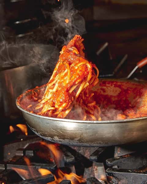 Steaming hot spaghetti being twirled in a skillet with rich marinara sauce over an open flame, capturing the bold aroma and heat of freshly made Italian pasta in a restaurant kitchen setting.