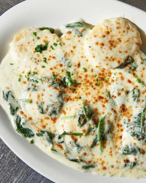 Cheese-stuffed ravioli smothered in a rich garlic cream sauce with sautéed spinach and topped with melted mozzarella and fresh parsley, served on a white ceramic plate.