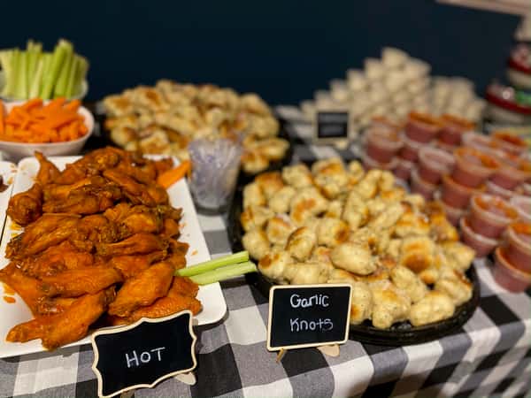 Buffet table topped with garlic knots, hot buffalo wings, celery and carrot sticks, and cups of marinara sauce, set on a black-and-white checkered cloth for a party-style appetizer spread.