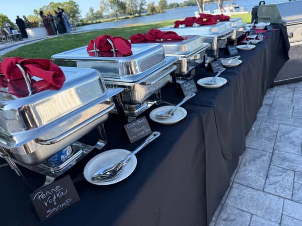 Outdoor buffet setup near a lakeside, showcasing stainless steel chafing dishes labeled with penne vodka with shrimp, gluten-free pasta, and other Italian-inspired entrees, perfect for weddings or upscale events.