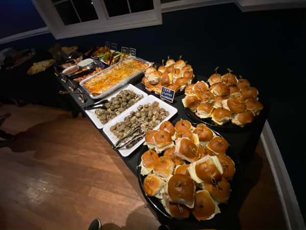 Buffet table at an indoor event featuring trays of crab-stuffed mushrooms, baked lasagna, assorted deli sliders on soft buns, and various hot entrees, ideal for casual catering or private parties.