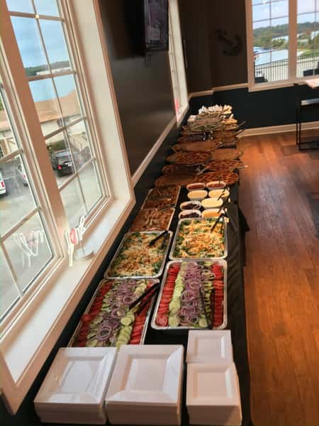 Long buffet table set near a bright window, displaying trays of fresh garden salads with cucumbers and onions, breaded appetizers, assorted pizzas, and dipping sauces for a casual event or corporate gathering.