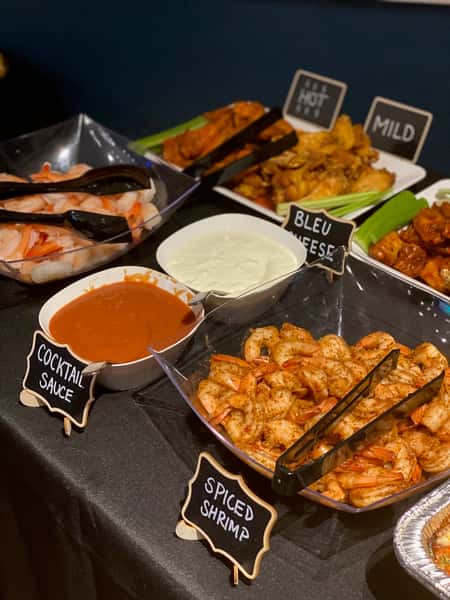 Buffet setup with chilled shrimp and spicy seasoned shrimp alongside cocktail sauce, blue cheese dip, and platters of mild and hot buffalo wings with celery sticks.