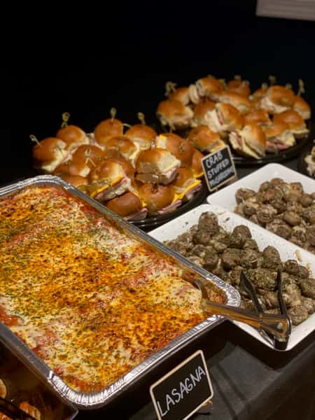 Close-up of a buffet spread with golden-baked lasagna, herb-seasoned crab-stuffed mushrooms, and deli sliders stacked on serving platters, arranged for a catered dinner or celebration.