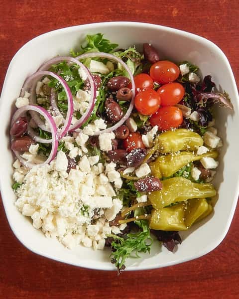 Fresh Greek salad in a white bowl featuring cherry tomatoes, red onions, kalamata olives, pepperoncini, crumbled feta cheese, and mixed greens over a red wooden table.