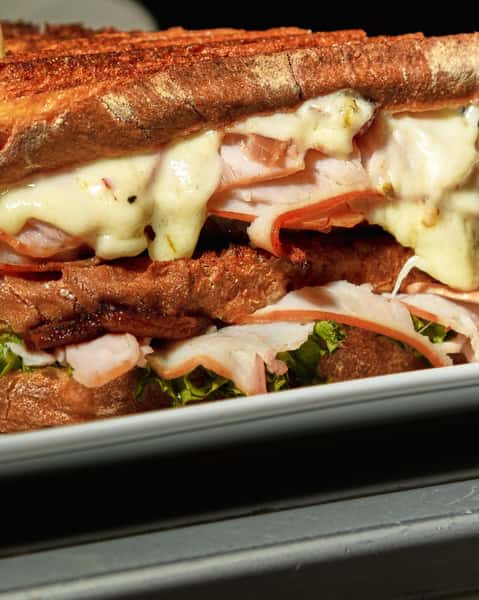 Close-up of a hot pressed sandwich loaded with sliced turkey, melted cheese, crispy bacon, and fresh lettuce on toasted artisan bread with golden grill marks.