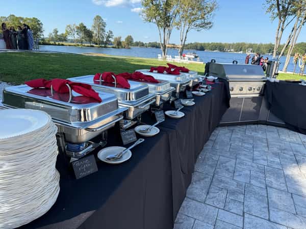 Chafing dishes lined along a black tablecloth at a waterfront event, labeled for pasta dishes like penne vodka and gluten-free options, accompanied by stacked white plates for outdoor catering.