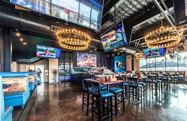 Spacious sports bar and restaurant interior with multiple TVs, high-top seating, modern industrial design, and chandelier lighting for game-day dining.