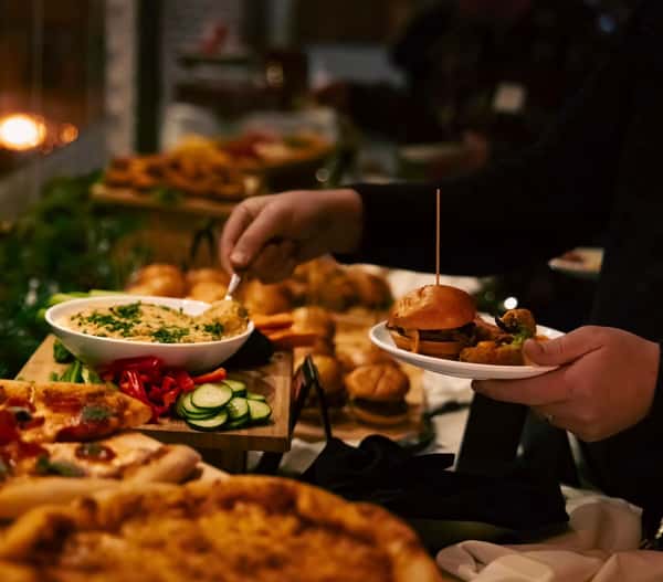 Guest serving gourmet sliders and appetizers from a catered buffet featuring pizzas, sandwiches, fresh vegetables, and event dining favorites.