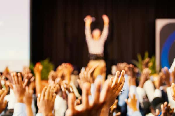 Engaged crowd raising hands during a live presentation or seminar, with a speaker blurred in the background on stage.