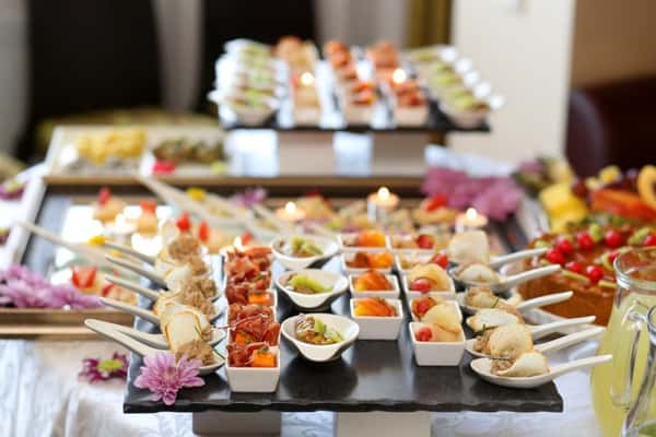 Fresh gourmet catering display with assorted bite-sized appetizers on elegant serving trays, including skewered canapés, bruschetta-style bites, and colorful hors d'oeuvres, arranged on a modern buffet table with candles and floral accents—ideal for upscale restaurant catering, event dining, or cocktail reception ambiance.