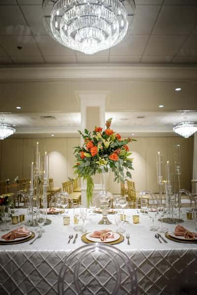Elegant banquet table setting with crystal chandelier overhead, tall floral centerpiece with orange roses and greenery, candlelight, and gold-accented place settings in a luxury event or wedding reception venue