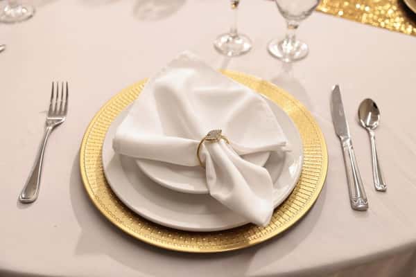 Elegant gold charger plate with a white dinner plate and neatly folded napkin in a decorative ring, paired with polished silverware on a formal wedding or upscale event table setting.