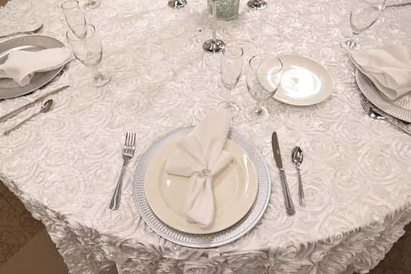 Elegant round banquet table with textured white tablecloth, silver charger plate, neatly folded white napkin in a decorative ring, polished silverware, and crystal glasses—refined restaurant-style table setting for a wedding reception, formal dinner, or upscale catered event.