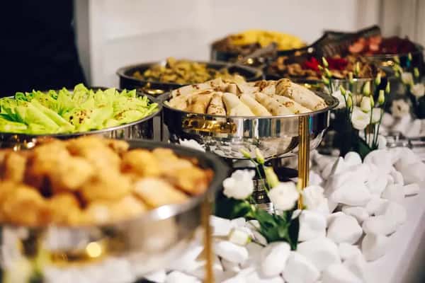 Elegant buffet catering setup with stainless steel chafing dishes filled with assorted hot foods, fresh salad, and appetizers on a decorated banquet table with white stones and floral accents.