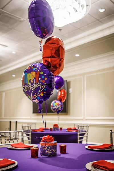 Birthday party event table setup with purple tablecloths, red napkins, and colorful helium balloons including a "Happy Birthday" balloon centerpiece—festive banquet hall décor for a catered birthday celebration or private party event.