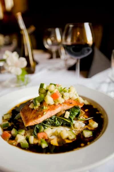 Pan-seared salmon served over sautéed greens with diced vegetable garnish and dark sauce, plated in an upscale restaurant dining setting with wine glasses