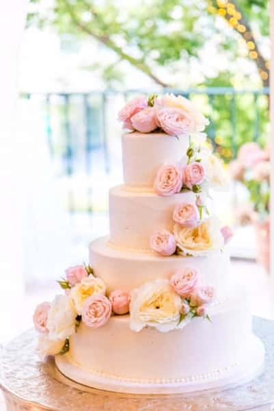 Three-tier wedding cake decorated with soft pink roses and white flowers, elegant dessert centerpiece at a romantic outdoor reception.
