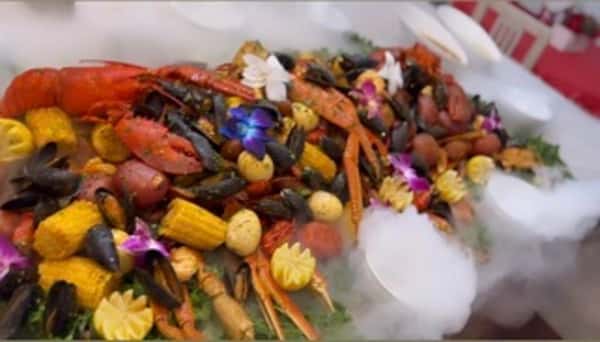 Seafood boil catering platter with lobster, mussels, corn, potatoes, and sausage garnished with edible flowers on a decorative buffet display.