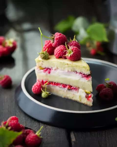 Slice of raspberry cream layer cake topped with fresh raspberries on a dark plate, elegant dessert presentation in a modern restaurant setting.