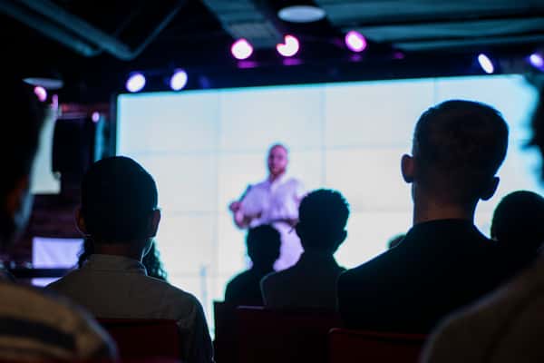 Attendees watching a speaker on stage with a large screen backdrop, capturing a modern conference or corporate event atmosphere.