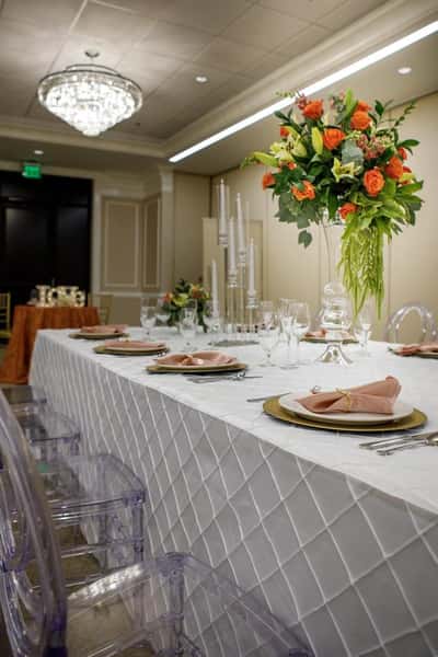 Luxury head table setup with white linens, gold-rimmed plates, crystal glassware, and tall floral centerpiece under a chandelier in a wedding venue.
