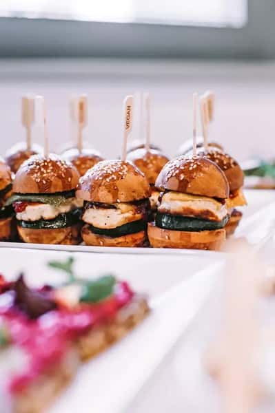Gourmet vegan slider burgers with sesame brioche buns, grilled vegetables, and plant-based patties served as elegant bite-sized appetizers on a catering platter.
