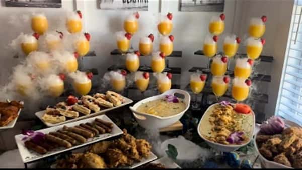 Upscale brunch buffet display with champagne-style orange mimosas garnished with strawberries, assorted appetizers, fried chicken, egg dishes, and gourmet finger foods arranged on an elegant catering table.