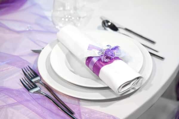 Elegant wedding place setting with white plates, silver cutlery, and a rolled napkin decorated with purple ribbon and floral accent on a formal banquet table