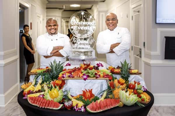 Professional chefs standing behind an elaborate fruit carving display with watermelon, pineapple, and tropical fruit arrangements at a catered event.