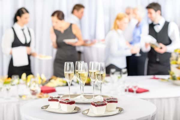 Champagne flutes and plated berry panna cotta desserts on an elegant catering table, with guests and waitstaff mingling in the background—refined restaurant-style event catering atmosphere with sparkling wine and gourmet dessert presentation.