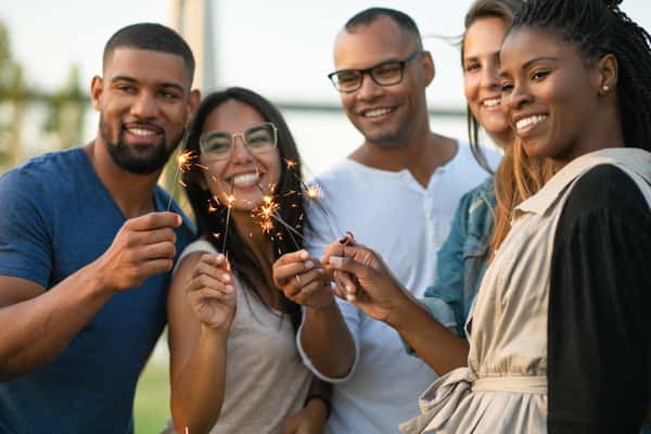 Smiling group of friends holding sparklers outdoors at sunset, festive celebration with joyful social vibe