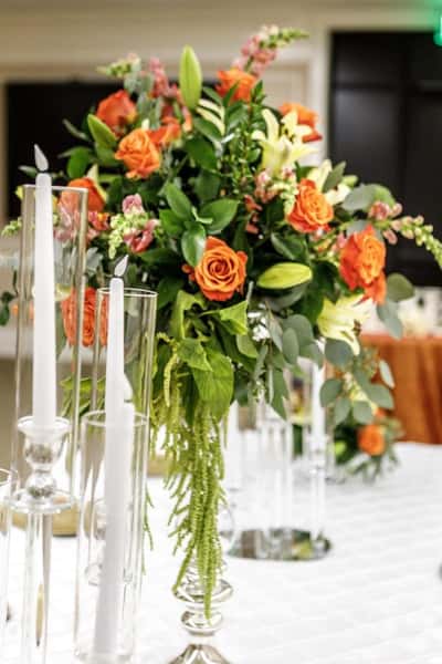 Tall floral centerpiece with orange roses, lilies, and greenery surrounded by glass candle holders on an upscale banquet dining table.
