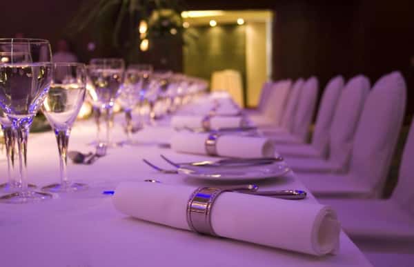 Long formal banquet table with neatly folded white napkins in silver rings, wine glasses, and soft ambient lighting—luxury restaurant or event venue table setting for fine dining and special occasions.