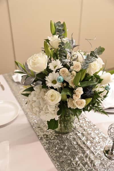 Elegant white floral centerpiece with roses, hydrangeas, daisies, and greenery in a glass vase on a decorated banquet table, creating a sophisticated wedding or event dining atmosphere
