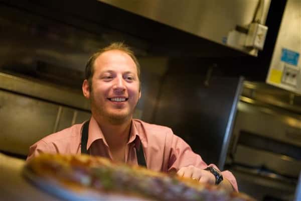 Man wearing an apron handing over a tray of pizza to someone