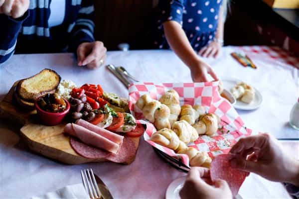 People eating from a basket of buns served with a dipping sauce and a wooden Charcuterie board on top of the table