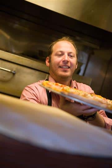 Man wearing an apron handing over a tray of pizza to someone