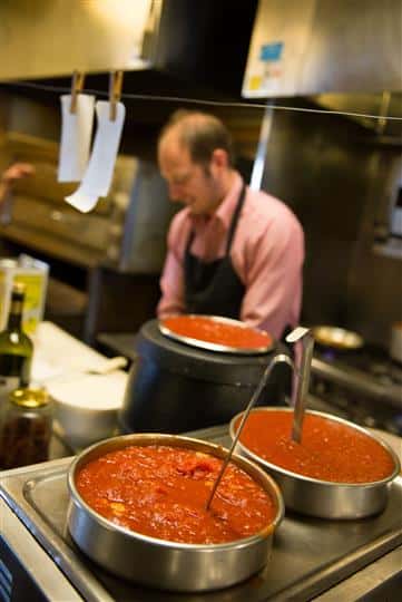 Photo of the kitchen with big pots of red sauce boiling and a man in the background preparing food