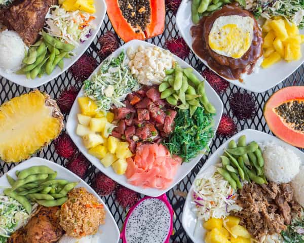 Overhead shot of assorted Hawaiian plate lunches including poke, loco moco, grilled meats, edamame, pineapple, rice, and tropical salads – vibrant and diverse island cuisine display.