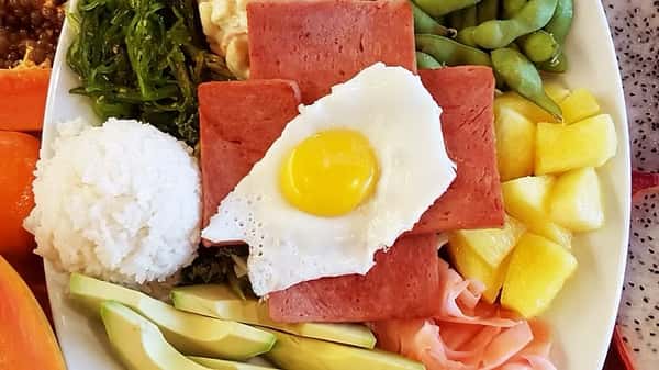 Hawaiian-style breakfast plate with thick seared Spam slices, a sunny-side-up egg, white rice, avocado, pickled ginger, pineapple, macaroni salad, seaweed, and edamame.