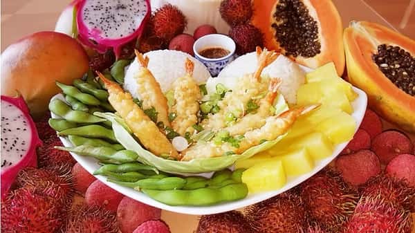 Golden shrimp tempura served with twin scoops of rice, edamame, pineapple chunks, and dipping sauce – surrounded by dragon fruit, mango, and papaya for a tropical island presentation.
