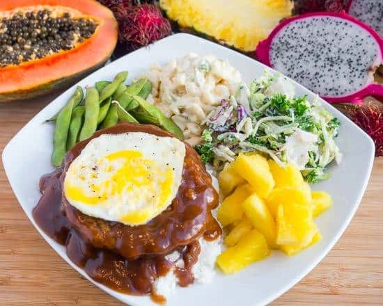 Traditional Hawaiian loco moco with a hamburger patty over rice, topped with a fried egg and rich brown gravy, served with edamame, macaroni salad, tropical slaw, and pineapple chunks.